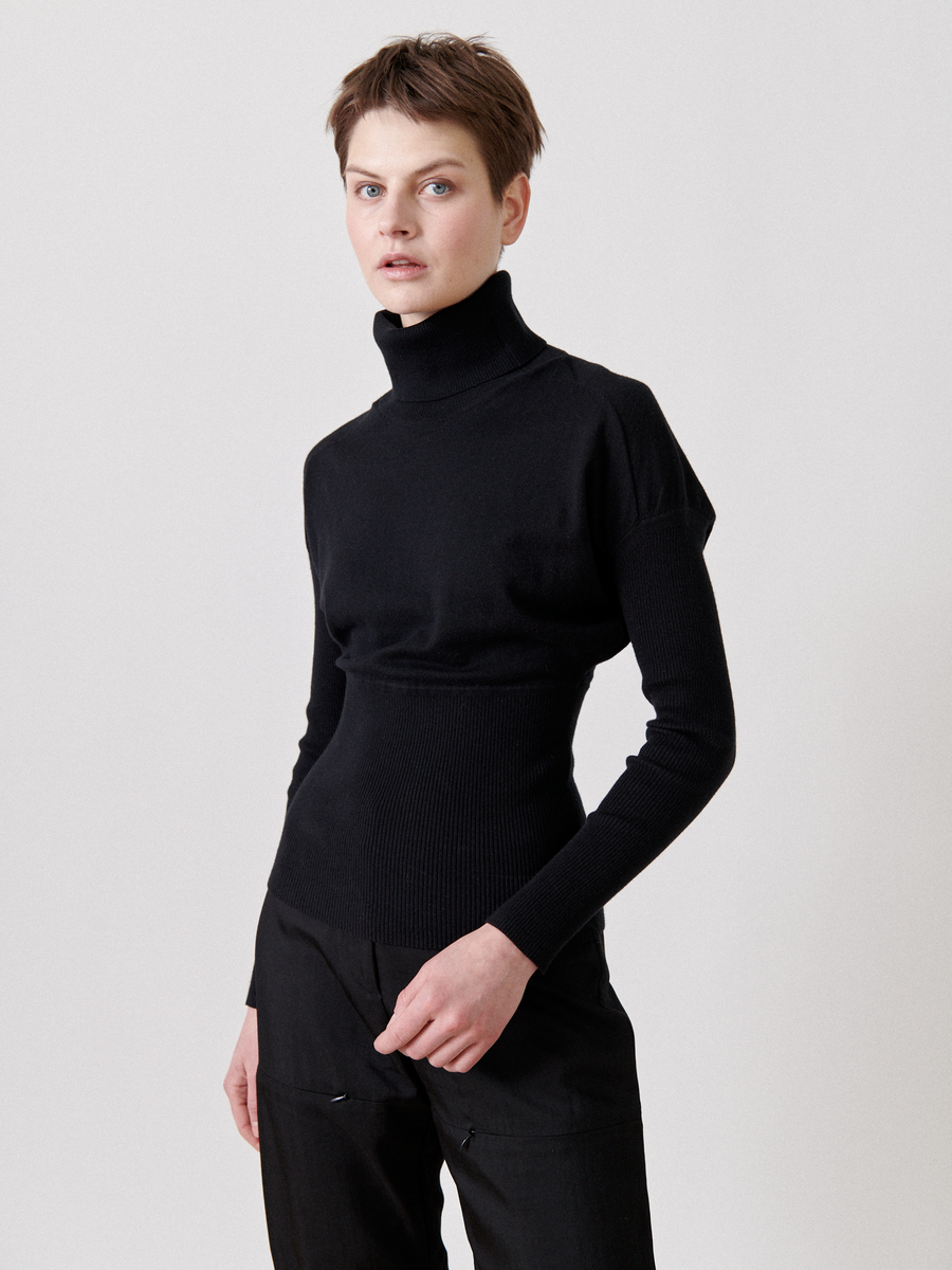 A person with short hair stands against a plain white background, wearing a black turtleneck sweater and black pants made of breathable luxury fabric. Their right hand rests on the side of their hip, and they look directly at the camera with a neutral expression. They are dressed in the Long-Sleeved Ama Rollneck by Zero + Maria Cornejo.