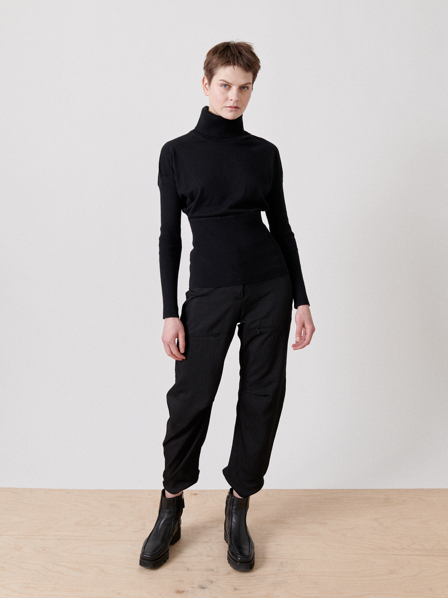 A person with short hair stands on a wooden floor against a plain white background. They are wearing a Long-Sleeved Ama Rollneck by Zero + Maria Cornejo made of breathable luxury fabric, black cargo pants, and black chunky boots. Their left hand rests at their side, and their right hand is slightly extended outward.