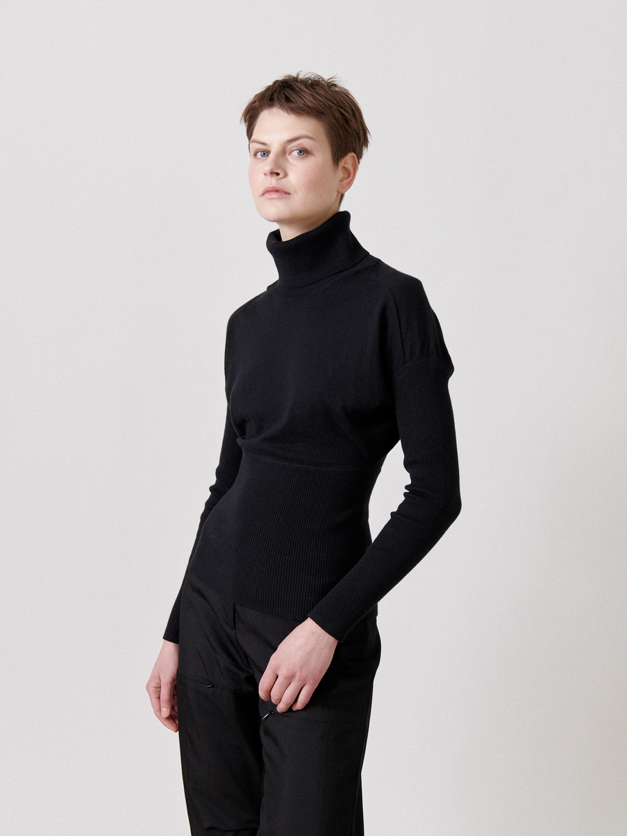 A person with short hair wearing a fitted, lightweight Zero + Maria Cornejo Long-Sleeved Ama Rollneck and black trousers poses against a plain, light-colored background. They are looking slightly off-camera with a calm expression, their left hand resting casually at their side.