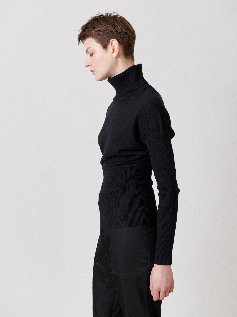 A person with short hair is standing in profile against a plain background. They are wearing a Zero + Maria Cornejo Long-Sleeved Ama Rollneck and black pants, their arms relaxed by their sides and eyes closed, presenting a calm, contemplative posture.