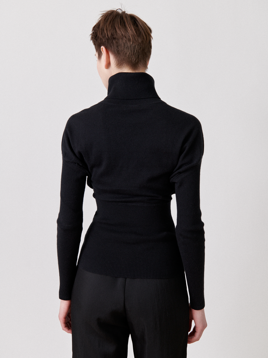 A person with short brown hair is standing facing away from the camera, wearing a black Zero + Maria Cornejo Long-Sleeved Ama Rollneck and black pants. The background is plain and light-colored.