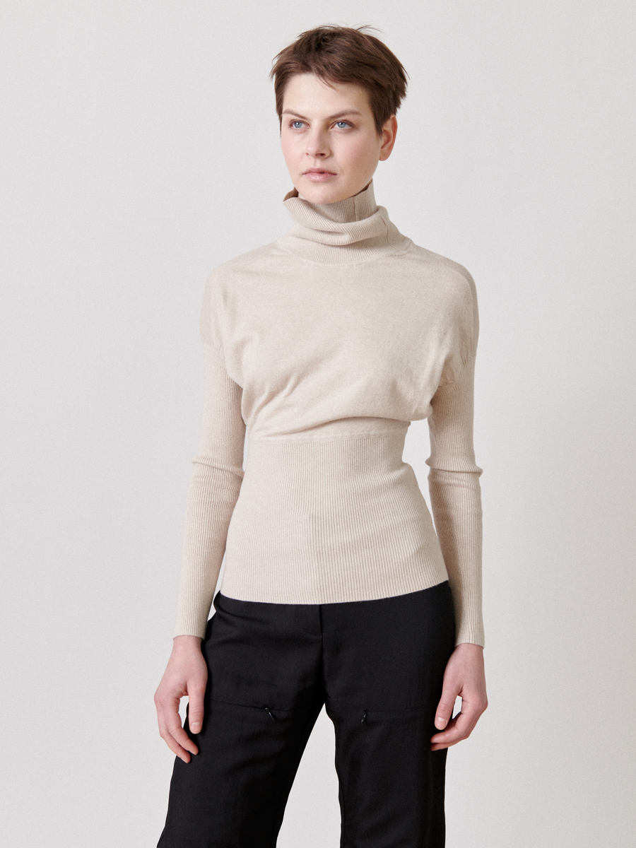 A person with short brown hair is wearing a beige Long-Sleeved Ama Rollneck from Zero + Maria Cornejo and black pants, standing against a plain white background. The person has a neutral expression and their hands are relaxed by their sides.