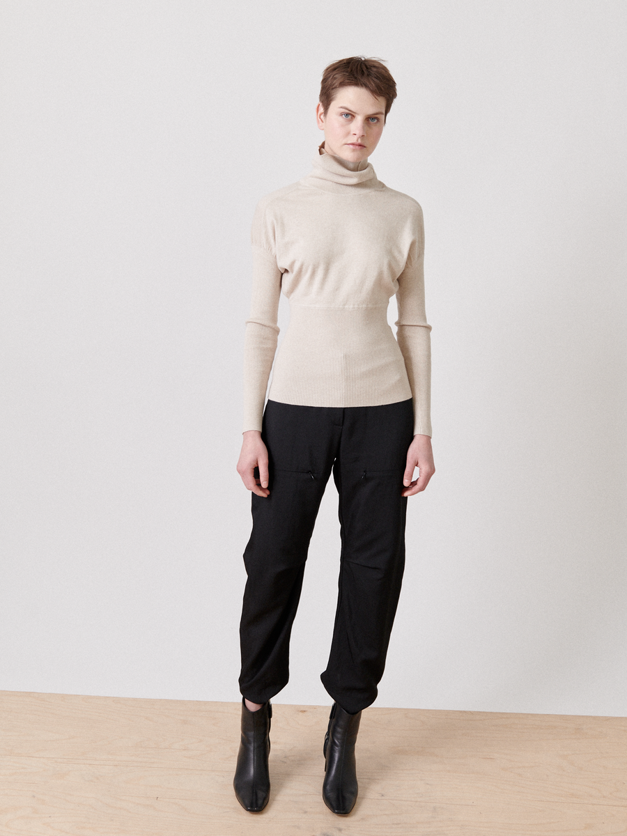 A person with short hair stands against a plain white background. They are wearing a light beige Zero + Maria Cornejo Long-Sleeved Ama Rollneck crafted from breathable luxury fabric and black cargo pants, paired with black ankle boots. Their expression is neutral, and their arms are relaxed by their sides.