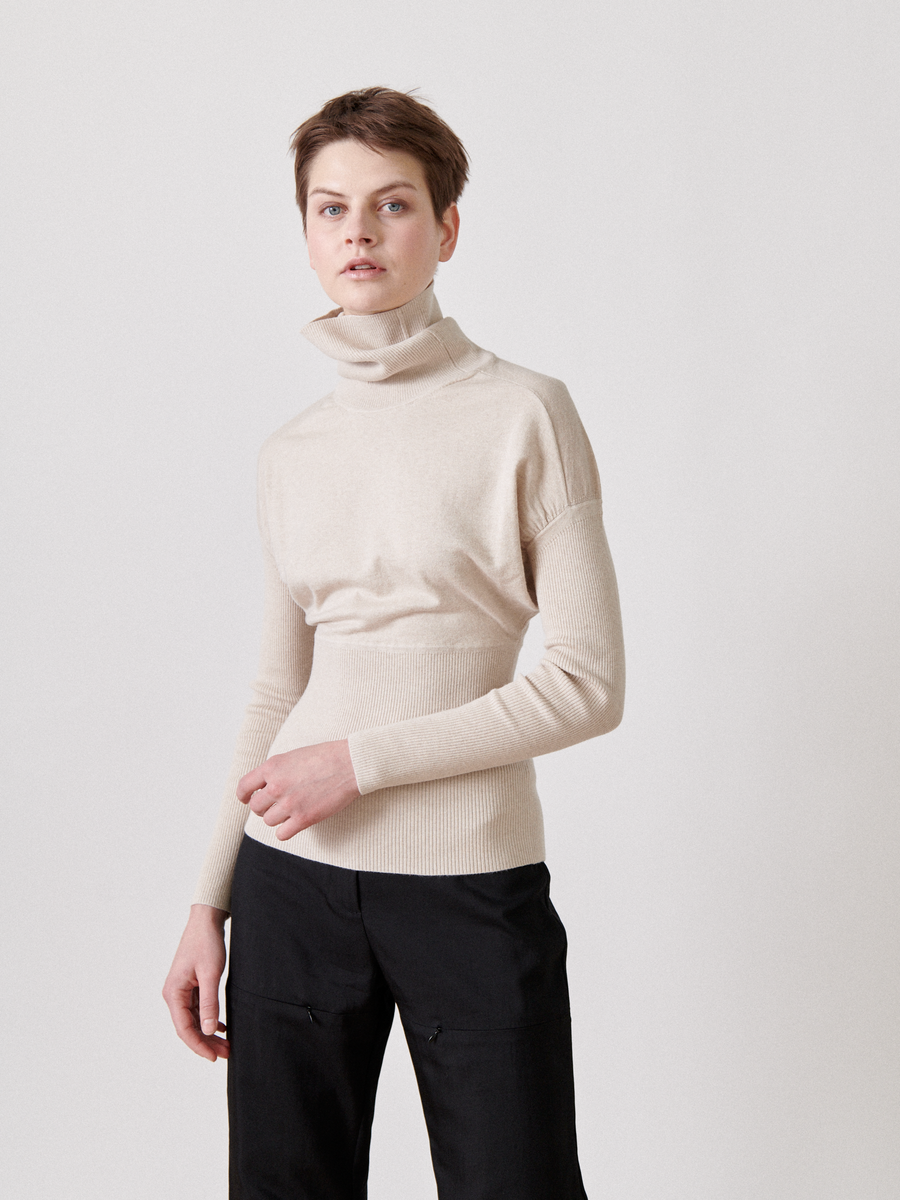 A person with short hair is wearing a cream-colored Zero + Maria Cornejo Long-Sleeved Ama Rollneck and black pants. They are standing against a plain, light-colored background, facing slightly to the left and looking directly at the camera with a neutral expression.
