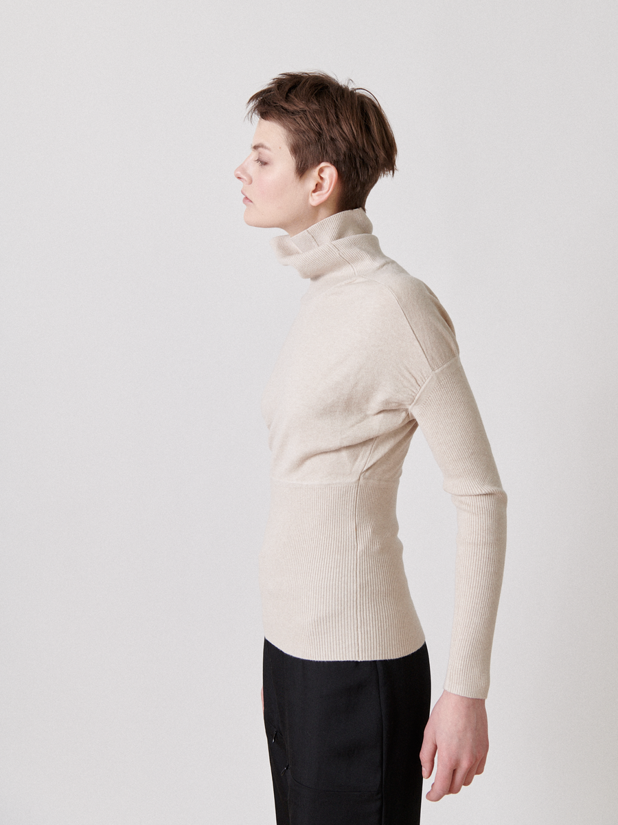 A person with short hair is shown in profile against a plain background. They are wearing a fitted, light beige Long-Sleeved Ama Rollneck by Zero + Maria Cornejo and dark pants. Their posture is upright, and their expression is neutral.