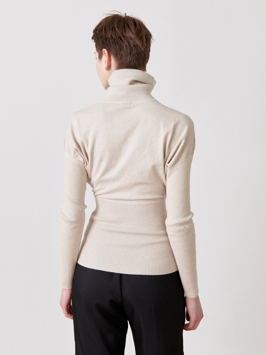 A person with short brown hair is seen from the back, wearing a light beige Zero + Maria Cornejo Long-Sleeved Ama Rollneck and black pants. The person stands against a plain, light-colored background.