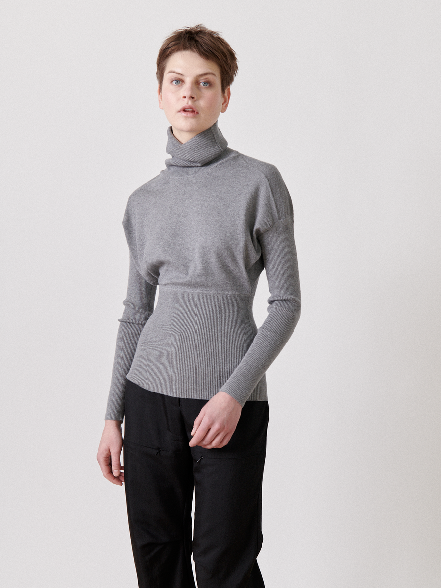 A person with short hair stands against a plain backdrop, wearing a gray Zero + Maria Cornejo Long-Sleeved Ama Rollneck and black pants. Their hands are down, and they have a neutral expression.