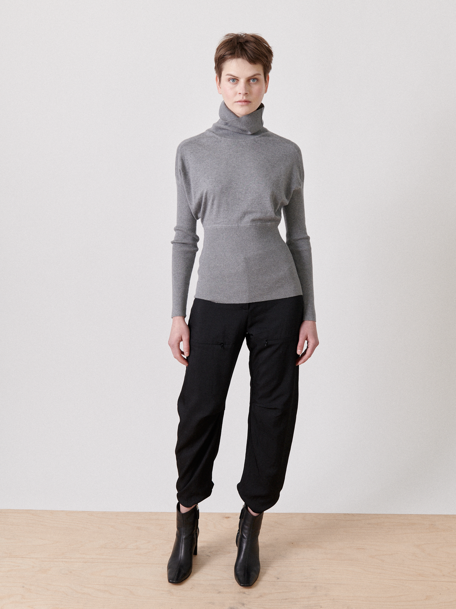 A person with short hair is standing against a plain light-colored background. They are wearing a gray silk-cashmere Long-Sleeved Ama Rollneck by Zero + Maria Cornejo, black pants with pockets, and black ankle boots. Their hands are by their sides and they are looking directly at the camera.
