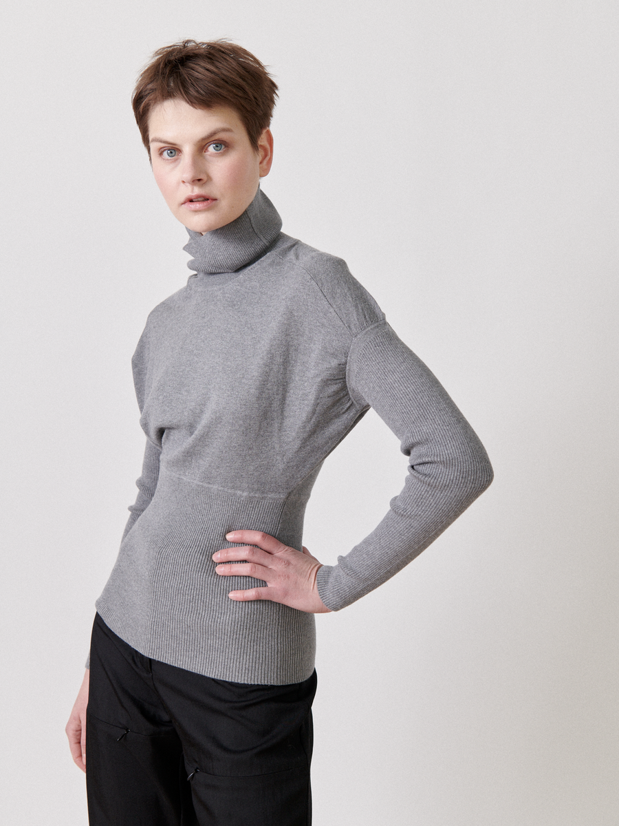 A person with short hair stands with one hand on their hip, wearing a fitted grey Long-Sleeved Ama Rollneck by Zero + Maria Cornejo and black pants. The background is plain and white. They look directly at the camera with a neutral expression.