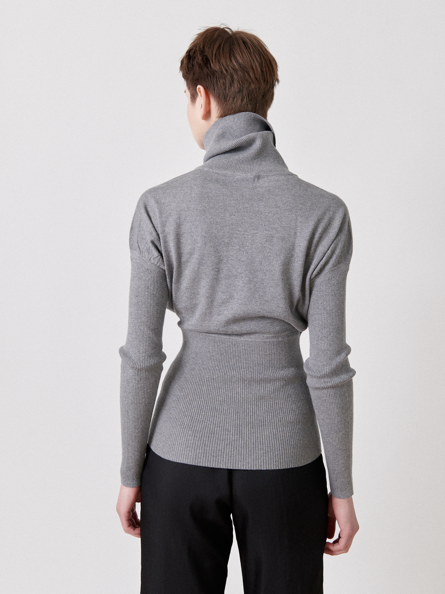 A person with short brown hair is seen from behind, wearing a grey Zero + Maria Cornejo Long-Sleeved Ama Rollneck and black pants. The sweater has a snug fit around the waist and ribbed detailing on the waistband and cuffs, exuding an air of breathable luxury fabric. The background is plain white.