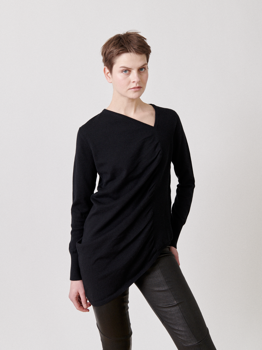 A person with short hair stands against a plain background, wearing the Off Kilter Tunic by Zero + Maria Cornejo with ruched detailing and black leather pants. They are gazing directly at the camera with a neutral expression and one hand resting on their hip.