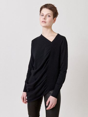 A person with short hair is wearing a black, asymmetrical knit sweater called the Off Kilter Tunic by Zero + Maria Cornejo, paired with black pants. They're standing against a plain, light-colored background and looking slightly upward and to the side.