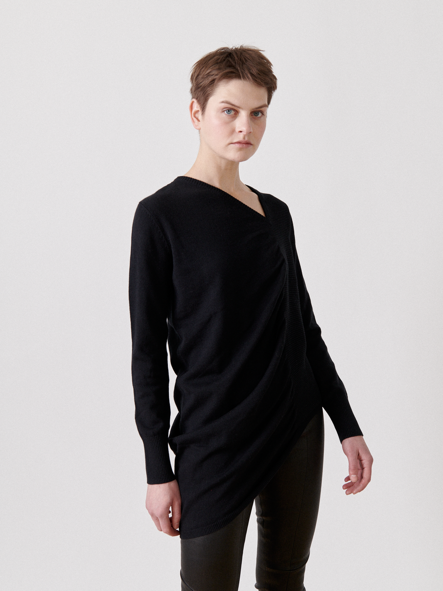 A person with short hair stands against a plain white background, wearing the Off Kilter Tunic by Zero + Maria Cornejo with an asymmetrical silhouette and dark pants. The top features delicate ruched detailing, creating a unique style. They have a neutral expression and their posture is relaxed.
