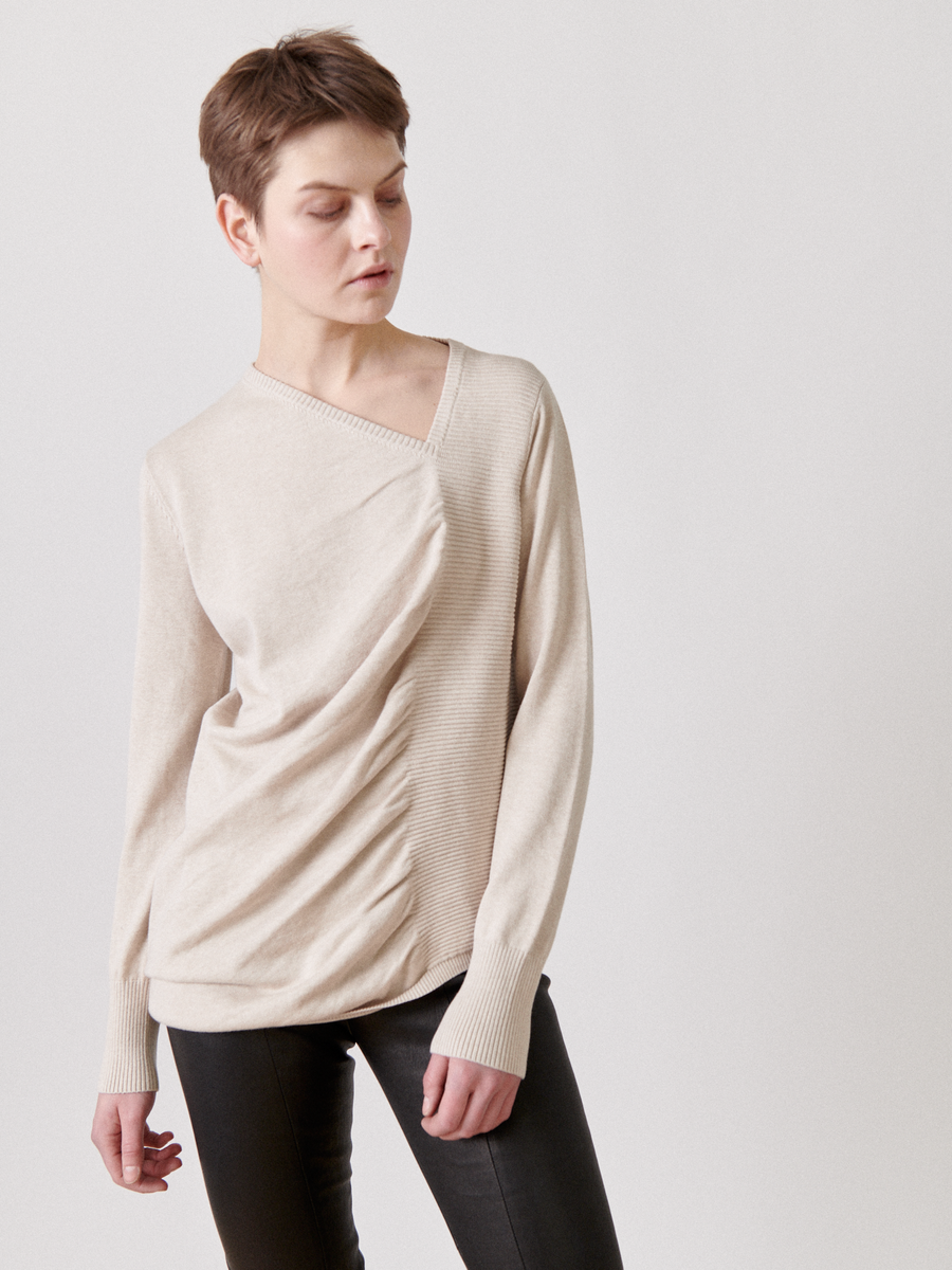 A person with short hair stands against a plain background. They are wearing a light beige, long-sleeved V-neck Off Kilter Tunic with subtle ruched detailing by Zero + Maria Cornejo and black pants. The individual's eyes are closed, and their head is tilted slightly to one side.