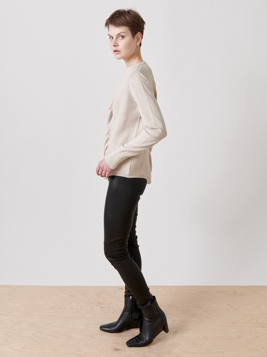 A person with short hair, wearing a beige long-sleeve Off Kilter Tunic by Zero + Maria Cornejo featuring ruched detailing, black leather pants, and black ankle boots stands sideways on a light wooden floor against a plain white background. Their hands rest at their sides.