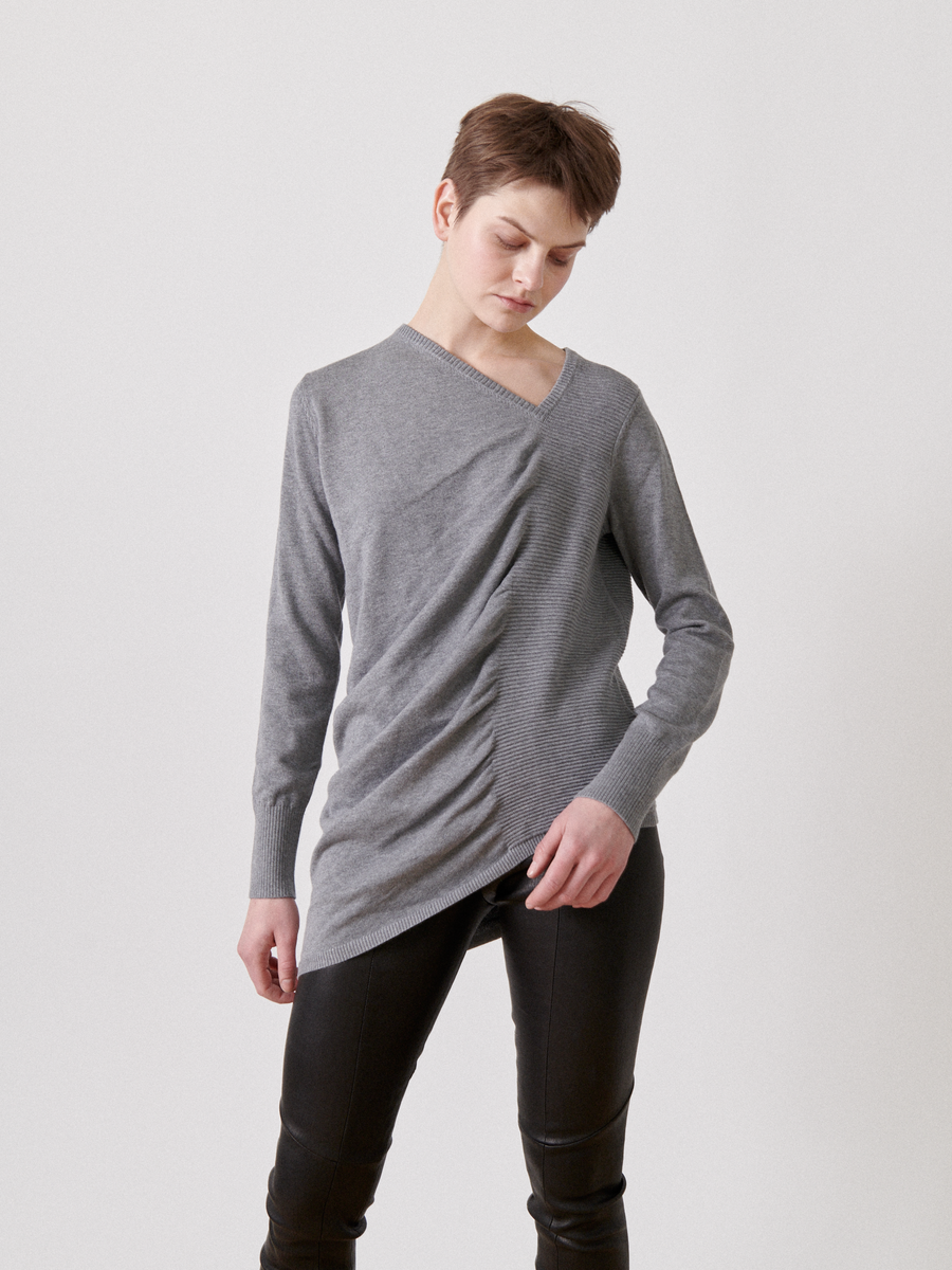 A person with short hair is wearing a stylish, asymmetrical grey sweater featuring ruched detailing on one side and black pants. They stand in a relaxed pose against a plain white background, with their eyes directed downward, wearing the Off Kilter Tunic from Zero + Maria Cornejo.