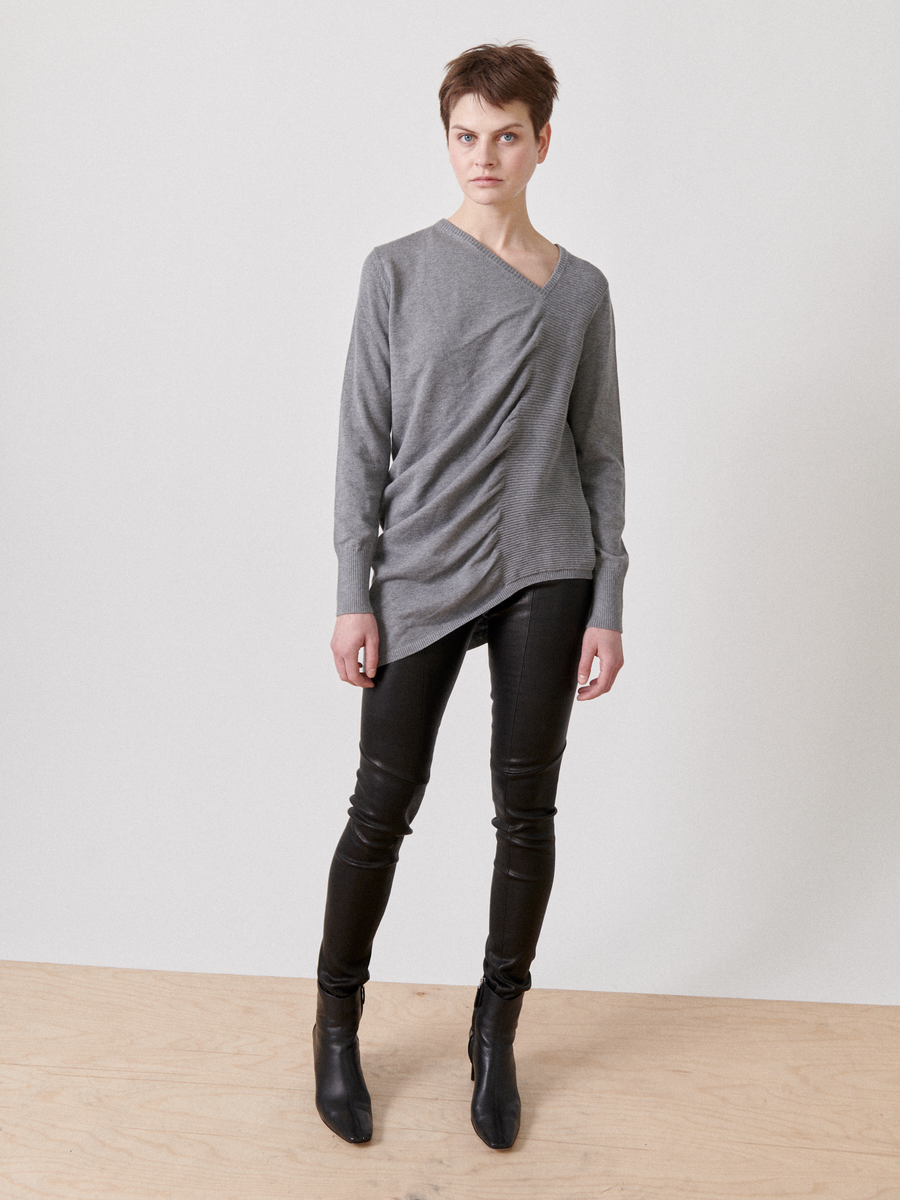A person with short hair stands against a plain white background. They are wearing an Off Kilter Tunic by Zero + Maria Cornejo with an asymmetrical silhouette and ruched detailing, paired with black leather pants and black ankle boots. Their hands are relaxed at their sides as they look directly at the camera.