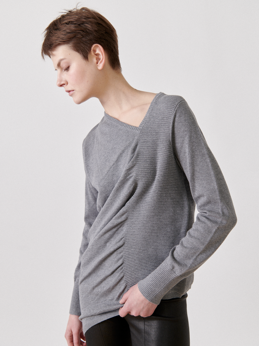 A person with short hair is wearing a gray, asymmetrical Off Kilter Tunic by Zero + Maria Cornejo with a unique pleated design on the front and ruched detailing. They are looking down with a contemplative expression. The background is plain white.