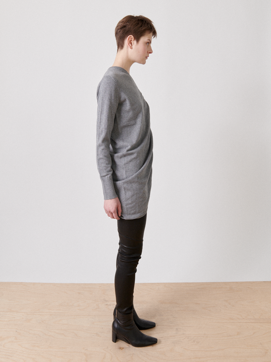 A person with short brown hair is standing in profile view against a plain white background. They are wearing the Off Kilter Tunic by Zero + Maria Cornejo, paired with black leather pants and black ankle boots.