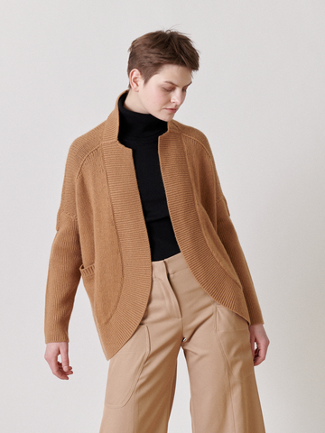 A person in a relaxed pose stands against a plain background, wearing the Long-Sleeved Rio Shrug from Zero + Maria Cornejo, paired with beige pants and a black turtleneck. They have short hair and are gazing slightly down and to the side.