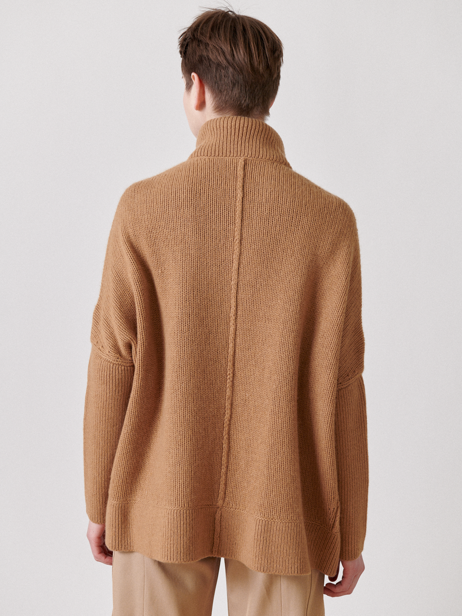 A person is seen from behind, wearing a cozy oversized tan turtleneck sweater with matching trousers. Crafted from recycled cashmere, the ribbed texture complements their short brown hair against a plain white background. The product may resemble Zero + Maria Cornejo's Long-Sleeved Rio Shrug - Final Sale.