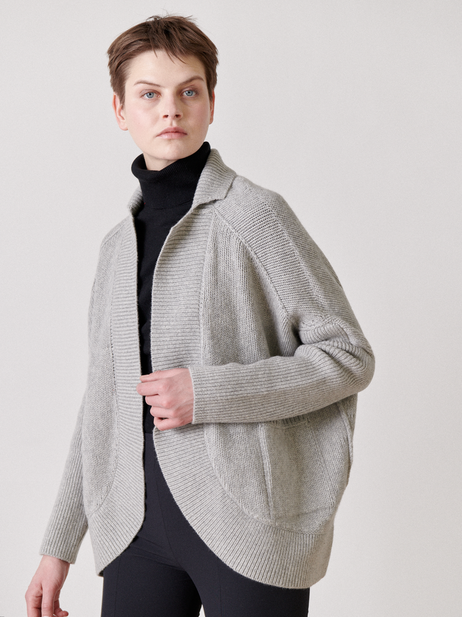 A person in a Long-Sleeved Rio Shrug by Zero + Maria Cornejo, made from recycled cashmere yarn, pairs it with a black turtleneck and pants. Their short hair enhances the minimalistic style against a neutral backdrop.