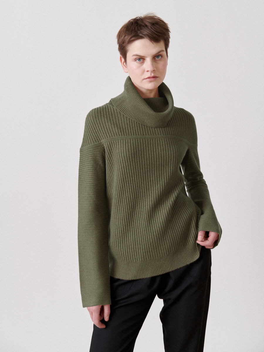 A person wearing a green Nian Rollneck by Zero + Maria Cornejo and black pants stands against a plain background, looking at the camera.