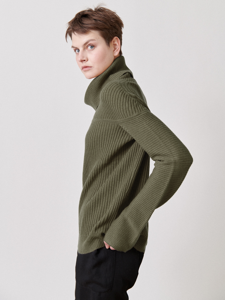 A person with short hair models the Zero + Maria Cornejo Nian Rollneck in olive green, made from recycled cashmere, paired with black pants. They stand in profile against a plain backdrop, gazing over their shoulder.