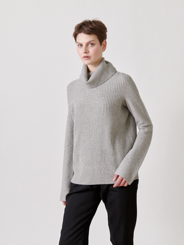 A short-haired individual in a light gray Nian Rollneck by Zero + Maria Cornejo and black pants stands against a plain white backdrop, looking sideways with a neutral expression.