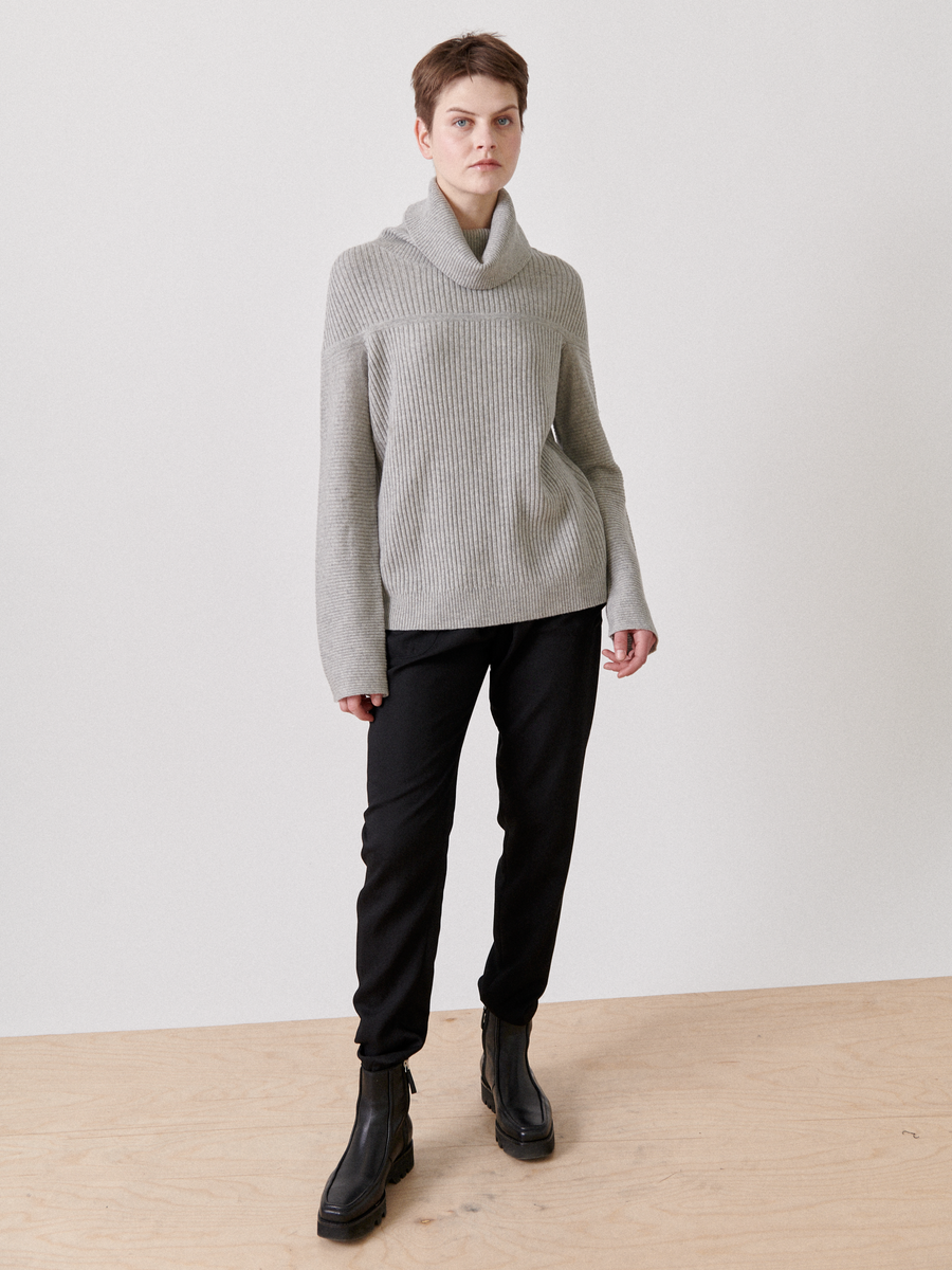 A person in a Zero + Maria Cornejo Nian Rollneck, black pants, and black ankle boots stands on a wooden floor against a white backdrop, gazing slightly to the side.