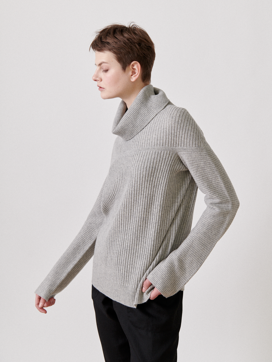 A person with short hair wears the Nian Rollneck in light gray from Zero + Maria Cornejo, paired with black pants, standing left against a plain white backdrop.