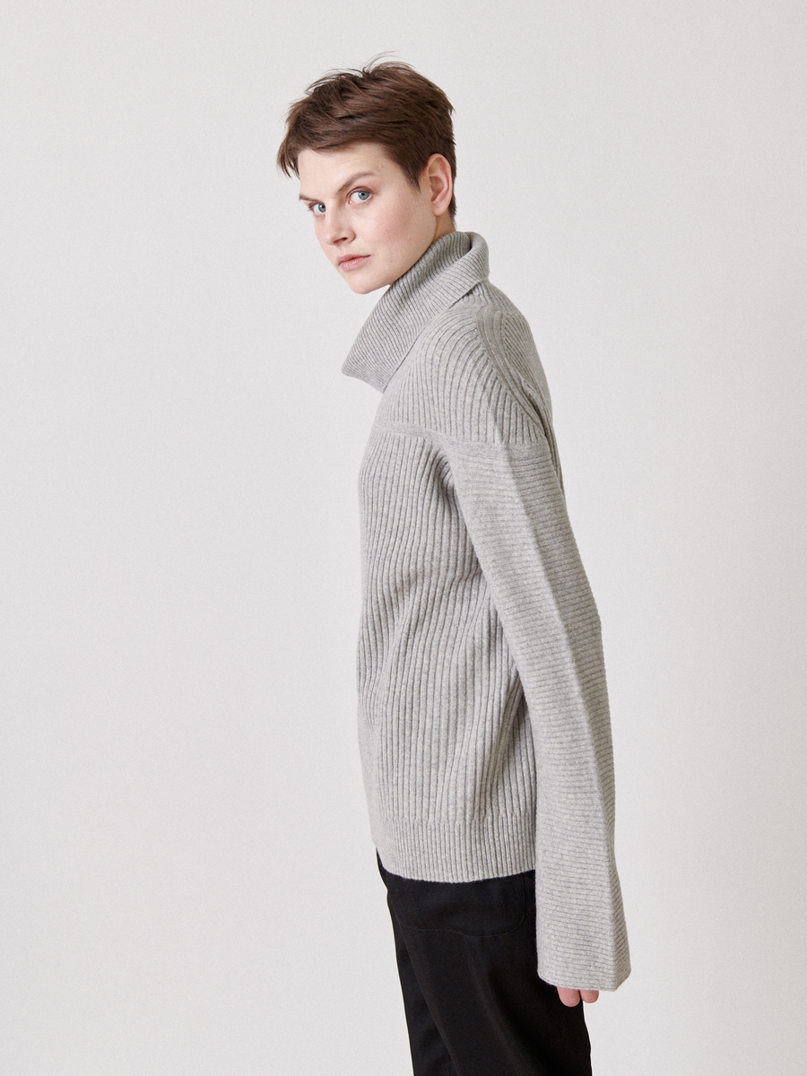 A person in the Nian Rollneck by Zero + Maria Cornejo, a gray ribbed turtleneck of recycled cashmere, pairs it with black pants. They stand against a plain white background, side-turned and facing forward. This item is marked as a final sale.