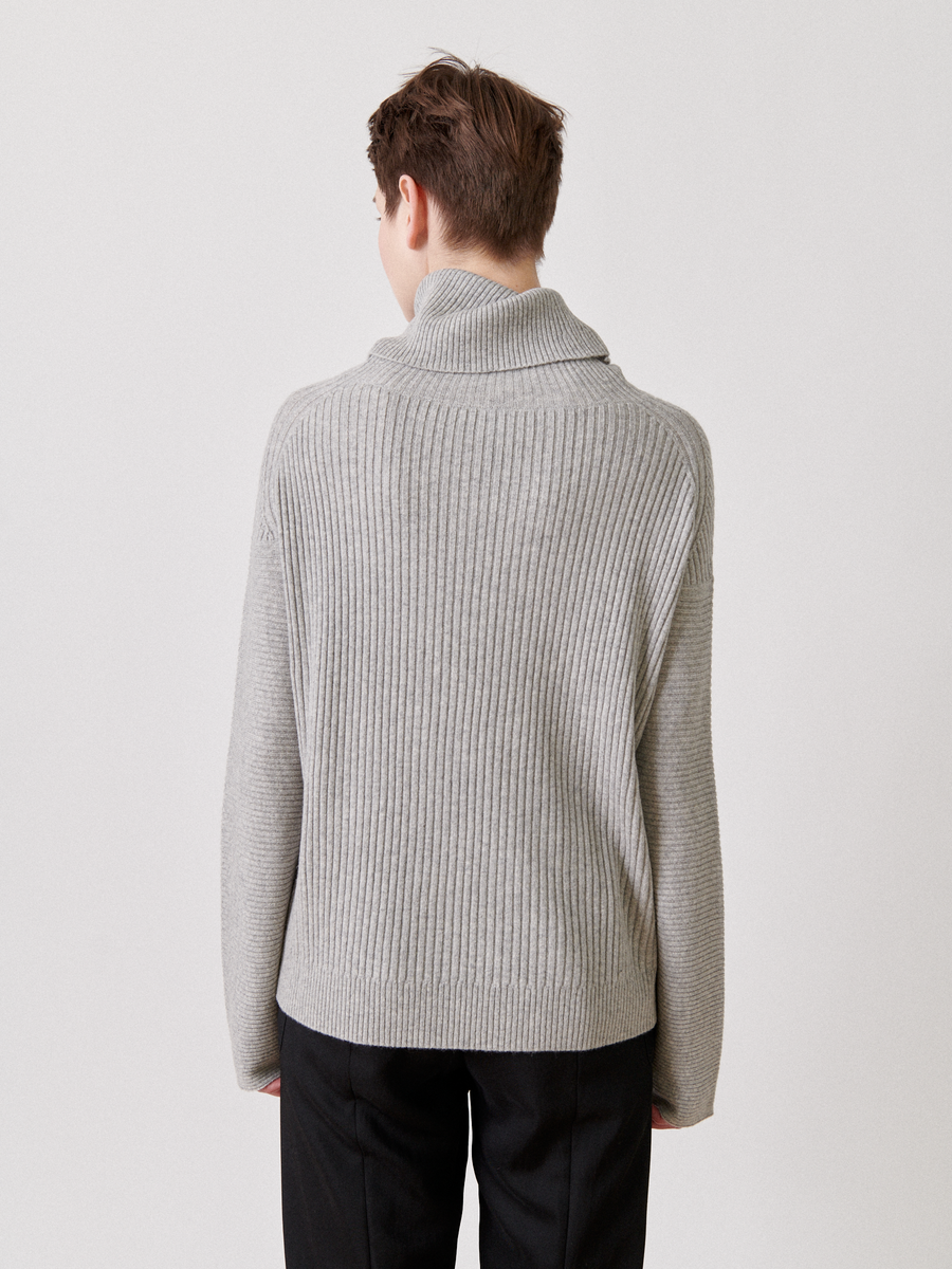 A person with short hair is seen from behind, wearing the Nian Rollneck by Zero + Maria Cornejo, a gray ribbed sweater made from recycled cashmere, paired with black pants against a plain light background.