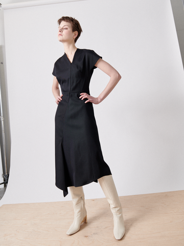 A person stands confidently with their hands on their hips, wearing a Silent Dress by Zero + Maria Cornejo and white knee-high boots. The background is a plain white backdrop set against a light wood floor. The person has short hair and is looking upward.