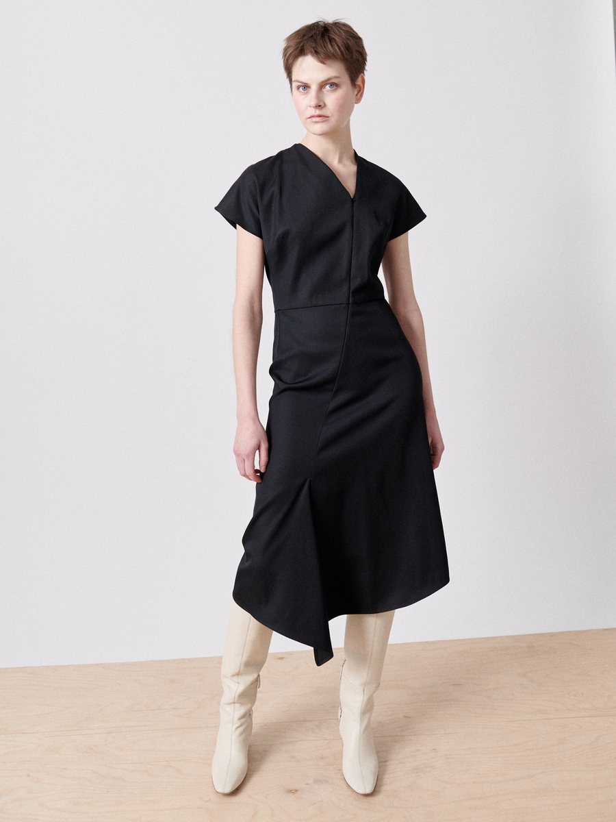 Person standing on a wooden floor against a plain white wall, wearing the Silent Dress by Zero + Maria Cornejo, made from FSC certified viscose yarns, paired with white knee-high boots. The person has short brown hair and adopts a confident pose, looking slightly off-camera.