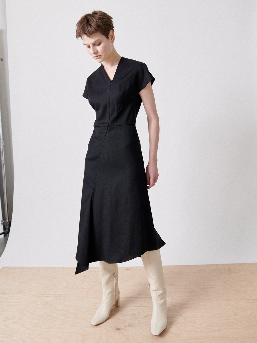 A person with short hair is standing against a plain backdrop, wearing the Zero + Maria Cornejo Silent Dress made from FSC certified viscose yarns. The black, short-sleeved midi dress features a draped skirt and is paired with white ankle boots with heels. They have a neutral expression and are looking slightly down and to the side.