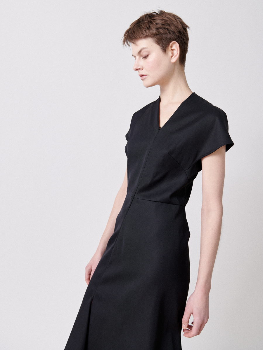 A person with short brown hair is wearing a black, asymmetrical v-neck Silent Dress by Zero + Maria Cornejo. They are slightly turned to one side, looking down with a neutral expression. The background is plain and light-colored, giving a clean and minimalistic look to the image.