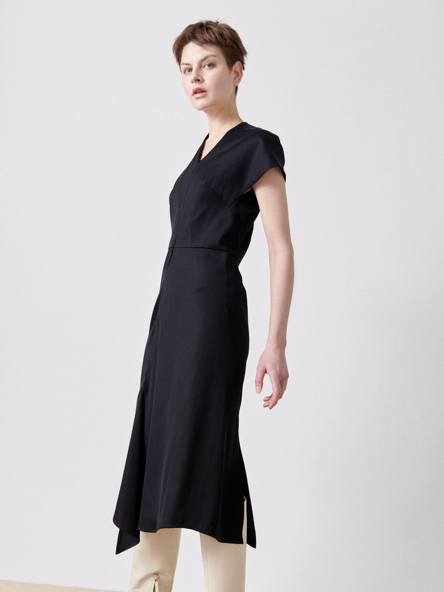 A person with short hair stands against a plain background, wearing an asymmetrical V-neck Silent Dress in black by Zero + Maria Cornejo that ends at the mid-calf, partially revealing white pants underneath. Their pose is slightly angled with one arm down and the other slightly raised.