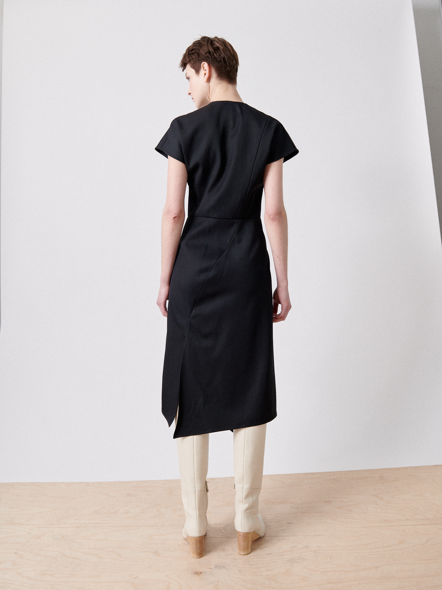 A person with short hair is standing on a wooden floor, facing a white wall. They are wearing an asymmetrical v-neck dress with a belt and beige knee-high boots. The Silent Dress by Zero + Maria Cornejo, made from FSC certified viscose yarns, features a side slit on the right and falls gracefully to the knees.