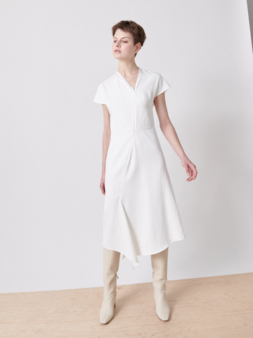 A person stands against a plain white background wearing Zero + Maria Cornejo's Silent Dress, featuring short sleeves and an asymmetrical v-neck. They have short hair, are donning tall light-colored boots, and pose with one arm slightly outstretched.