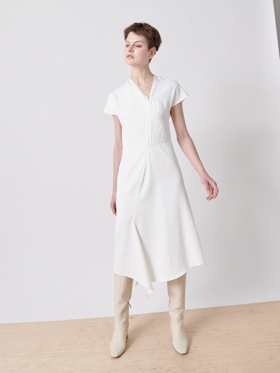 A person stands against a plain white background wearing Zero + Maria Cornejo's Silent Dress, featuring short sleeves and an asymmetrical v-neck. They have short hair, are donning tall light-colored boots, and pose with one arm slightly outstretched.