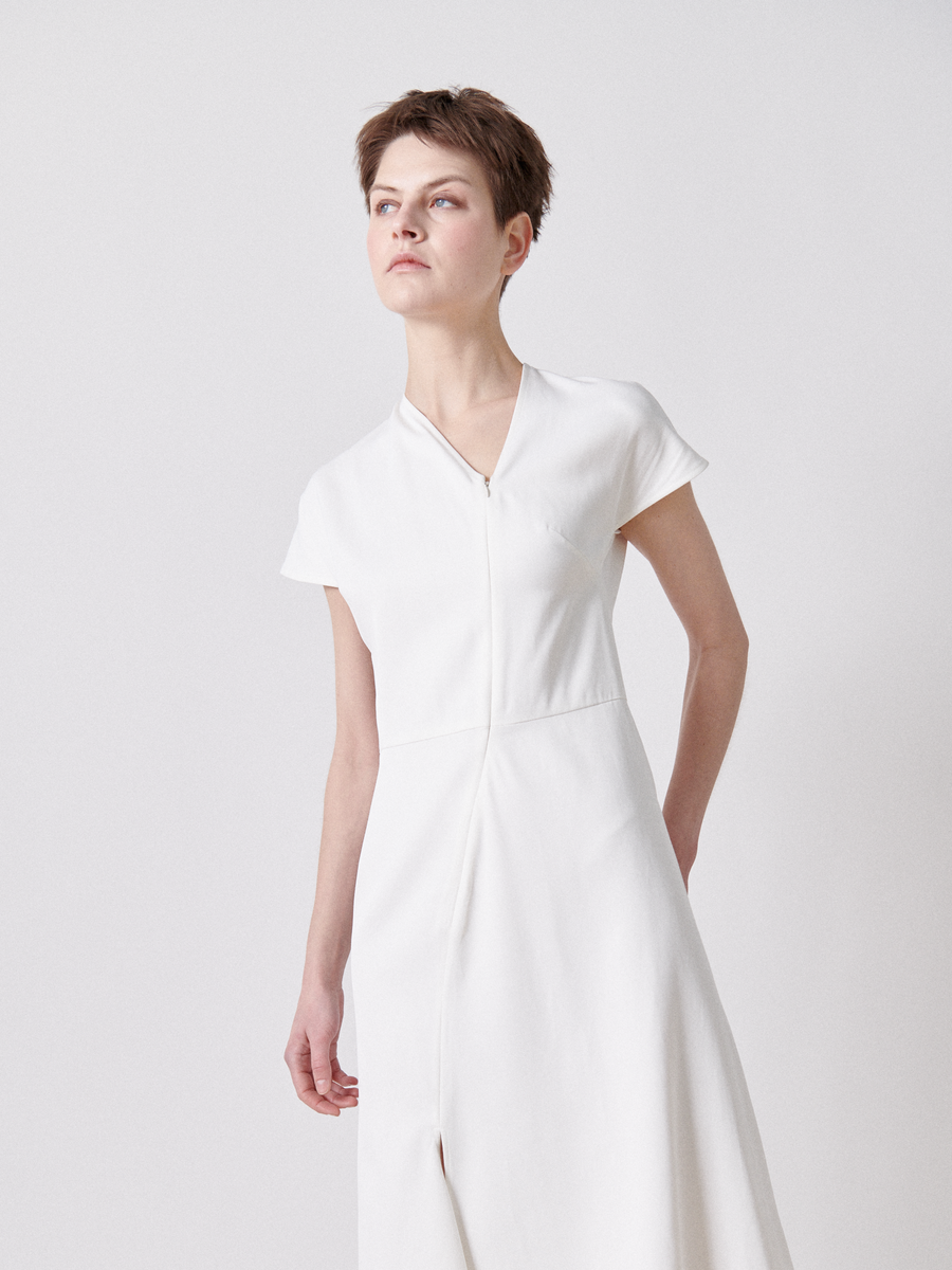 A person with short hair models the Silent Dress by Zero + Maria Cornejo, a final sale item. The dress is a simple yet elegant white twill design with an asymmetrical V-neckline and short sleeves. They stand against a plain background, gazing upwards.