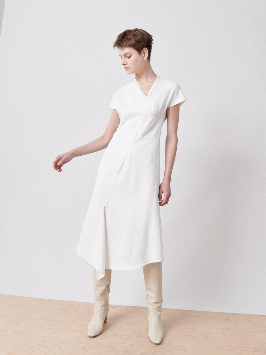 A person with short hair wears the Silent Dress by Zero + Maria Cornejo, featuring white short sleeves and beige knee-high boots, standing on a wooden floor against a plain white background, with one arm slightly bent and looking to the side.