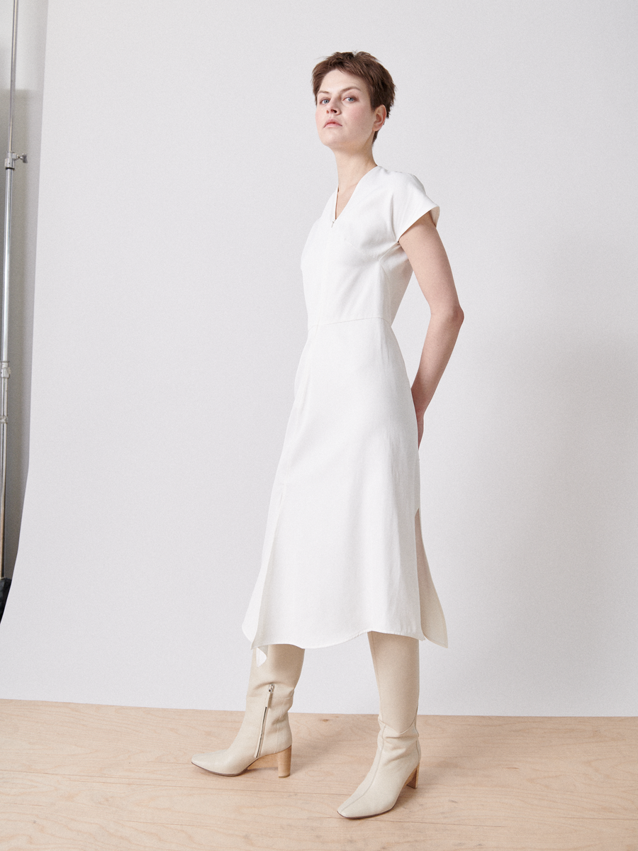 A person stands on a light wooden floor against a white backdrop, wearing the Silent Dress - Final Sale by Zero + Maria Cornejo, featuring short sleeves and an asymmetrical v-neck, paired with cream knee-high boots, gazing slightly to the side.