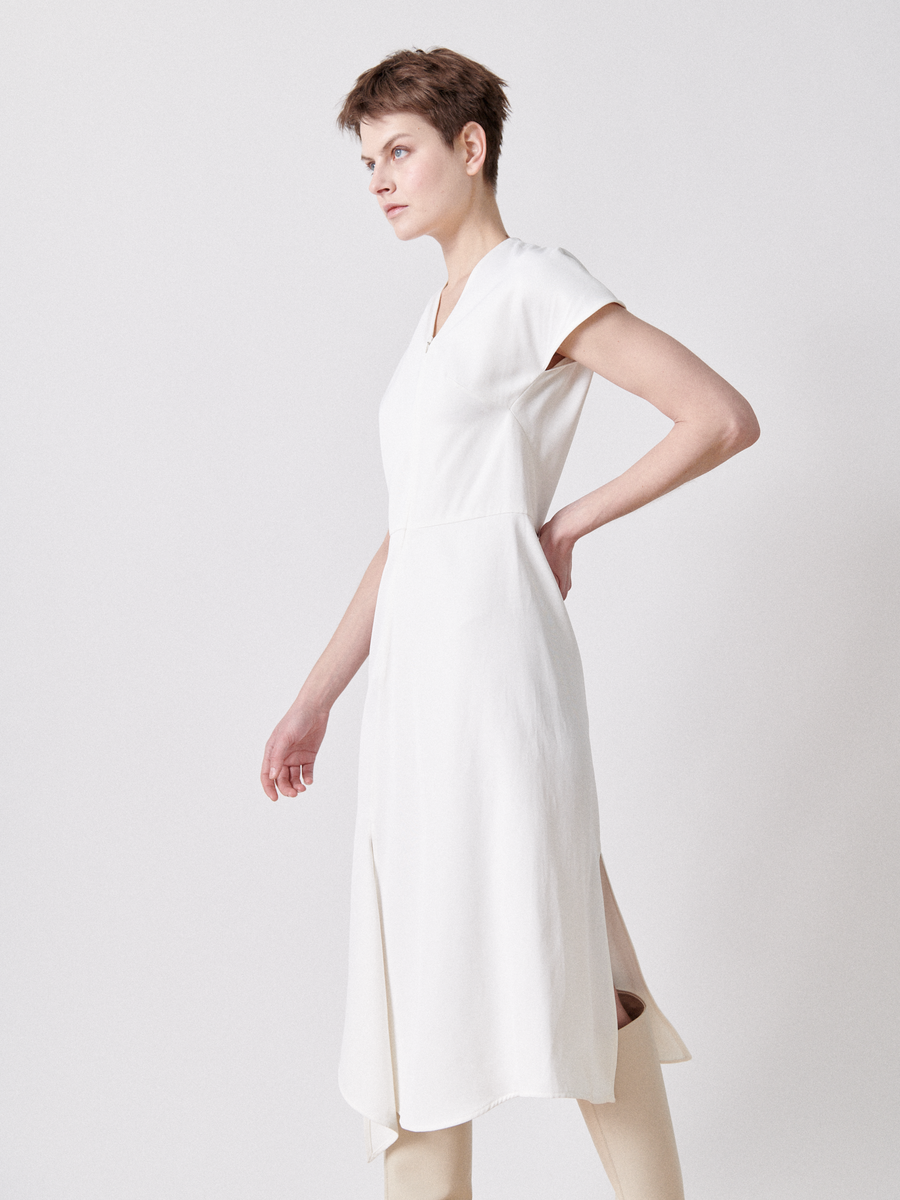 A person with short hair poses confidently in a Zero + Maria Cornejo Silent Dress - Final Sale. Featuring a sleek, short-sleeved style with an asymmetrical V-neck and side slit that reveals beige pants, they stand against a plain background in side profile, hand on hip.