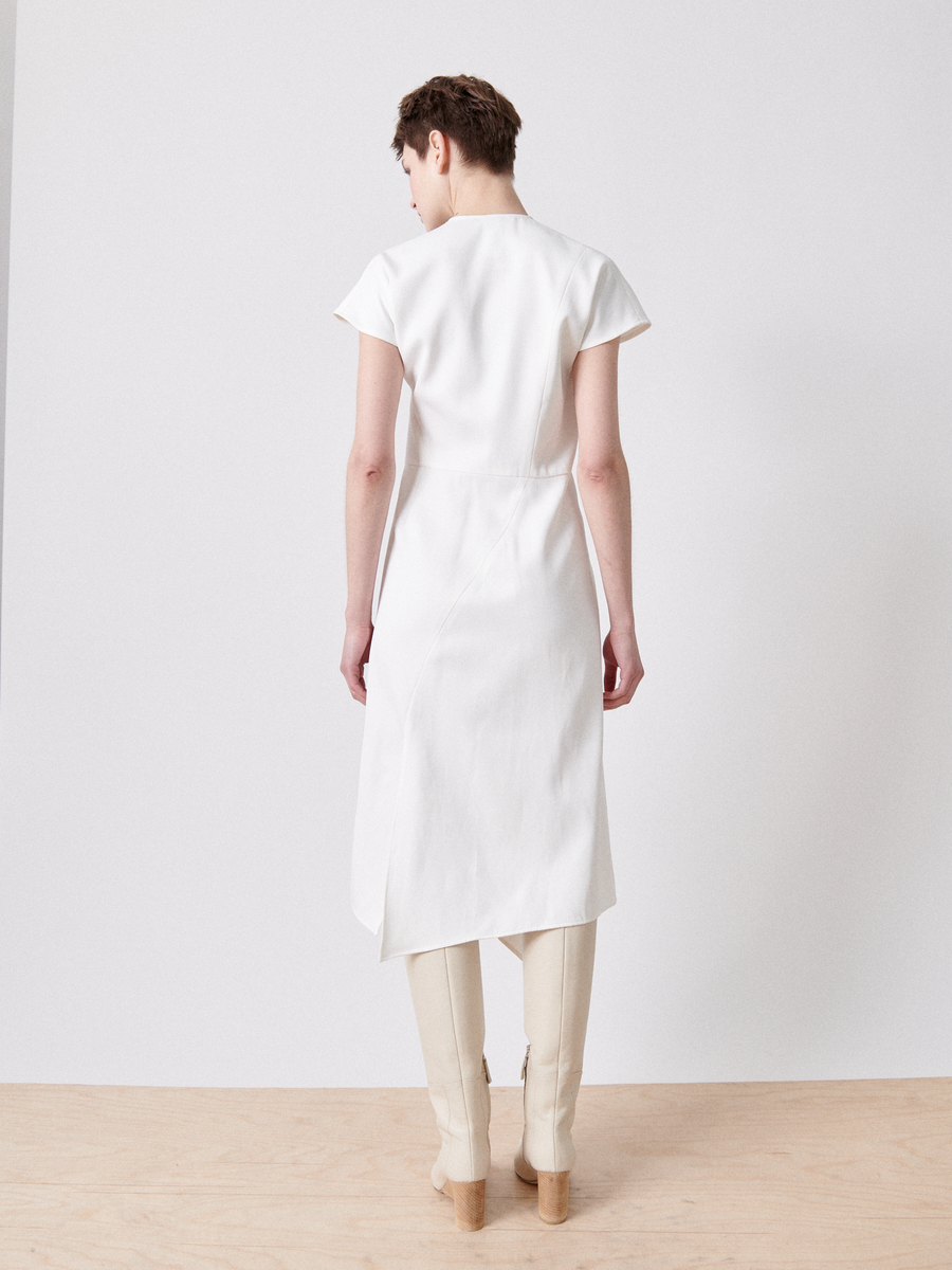 A person with short hair stands in a minimalistic room, wearing the Silent Dress by Zero + Maria Cornejo, an FSC-certified viscose dress with short sleeves and an asymmetrical hem. The light wood floor and white walls accentuate the elegance of the dress paired with light-colored tall boots.