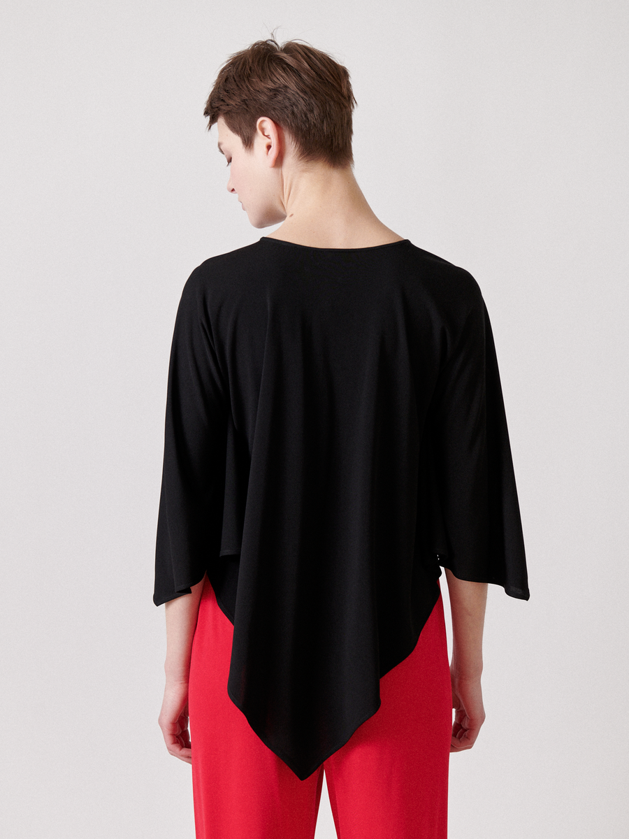 A person with short brown hair wears Zero + Maria Cornejo's Triangle Top, a black asymmetrical lightweight jersey, paired with vibrant red pants. The draped, flowing silhouette is viewed from the back against a neutral gray background.