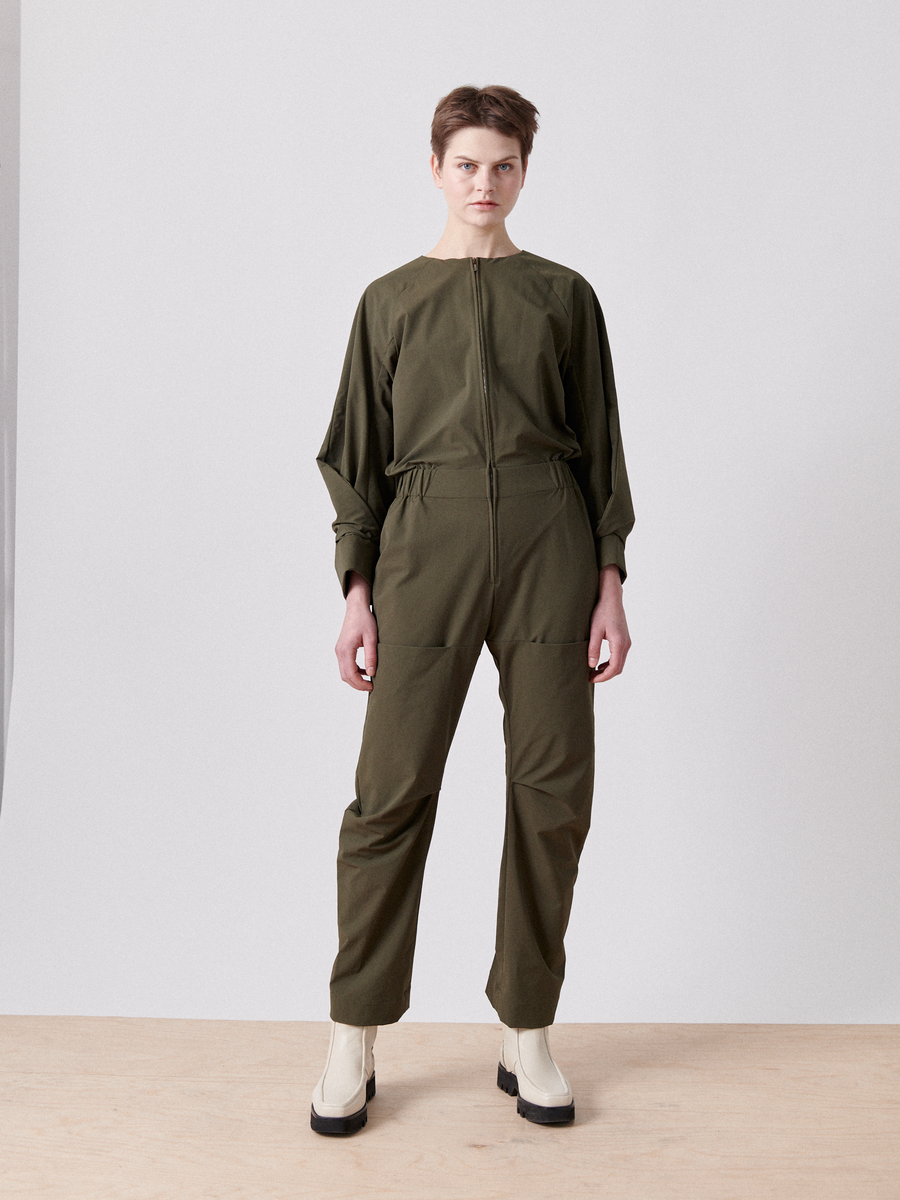 Against a light gray backdrop, a person wears the Biker Jumpsuit - Final Sale by Zero + Maria Cornejo in olive-green. It has an elastic-banded waist, front zipper, and loose sleeves. Complemented by cream boots, their short hair is simply styled over a light wooden floor.