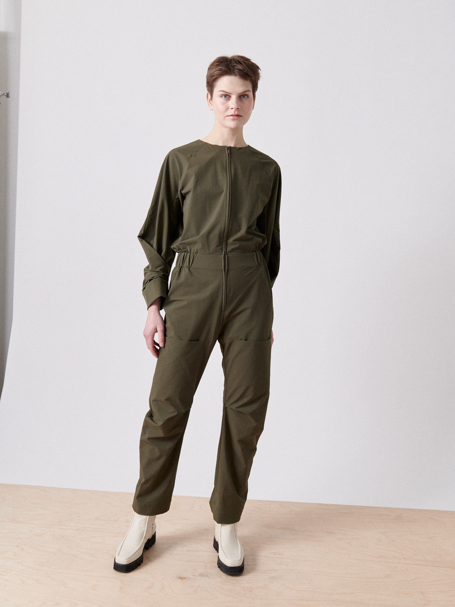 A person on a light wood floor wears the olive green Biker Jumpsuit by Zero + Maria Cornejo. With short hair, they pair it with white boots featuring black soles. A plain light gray wall in the background complements this minimalist look.