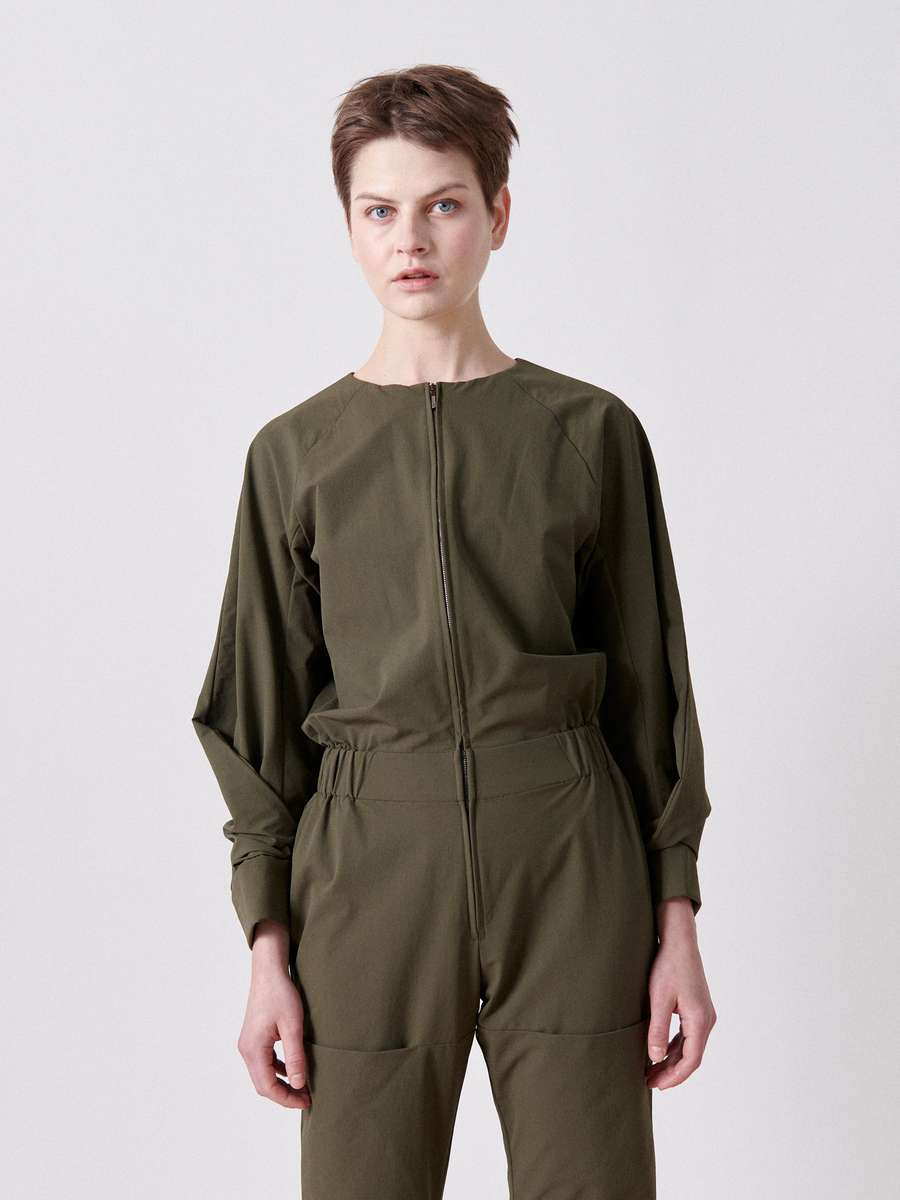 An individual with short hair wears the Biker Jumpsuit by Zero + Maria Cornejo. This sleek ensemble, in olive green technical stretch fabric, features a round neck, front zipper, and elastic banded waist against a plain light-colored background. (Final Sale).